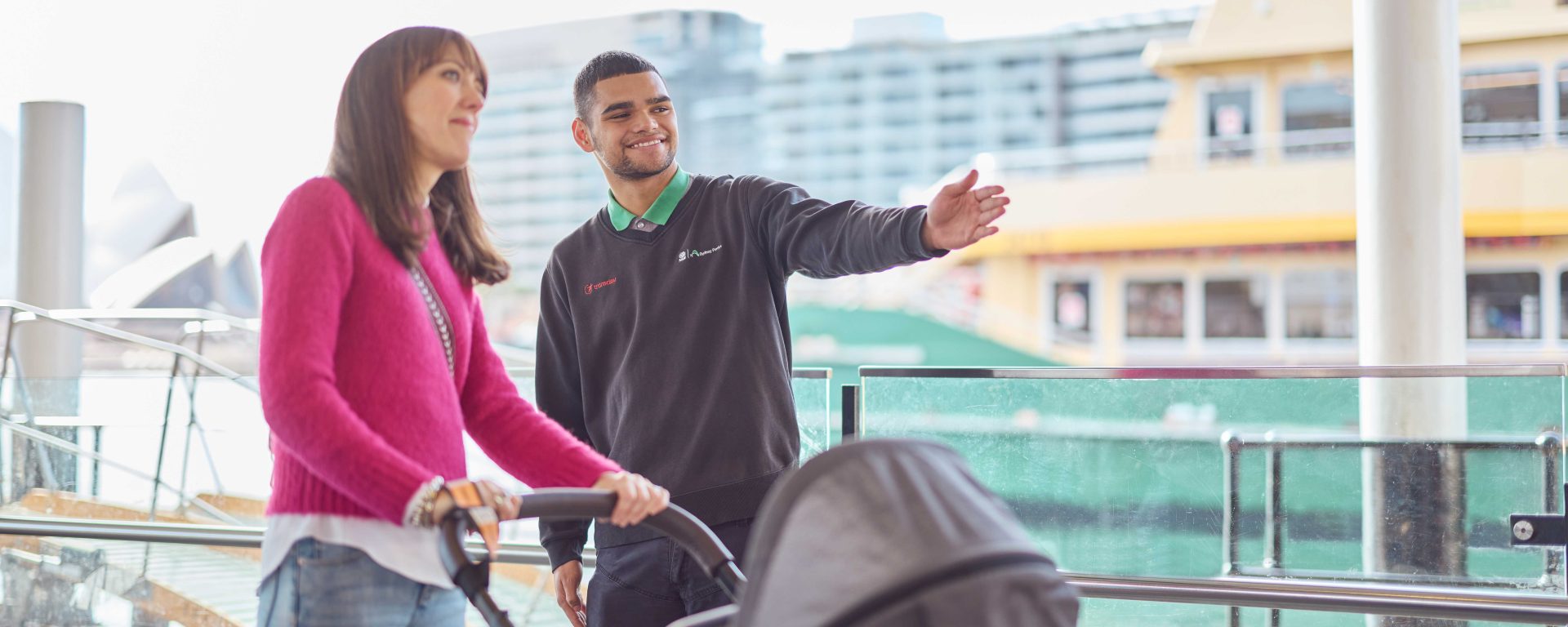 An employee of Transdev Sydney Ferries provides directions to a woman in a pink sweater with a pram. They are at Sydney's Circular Quay Wharf.