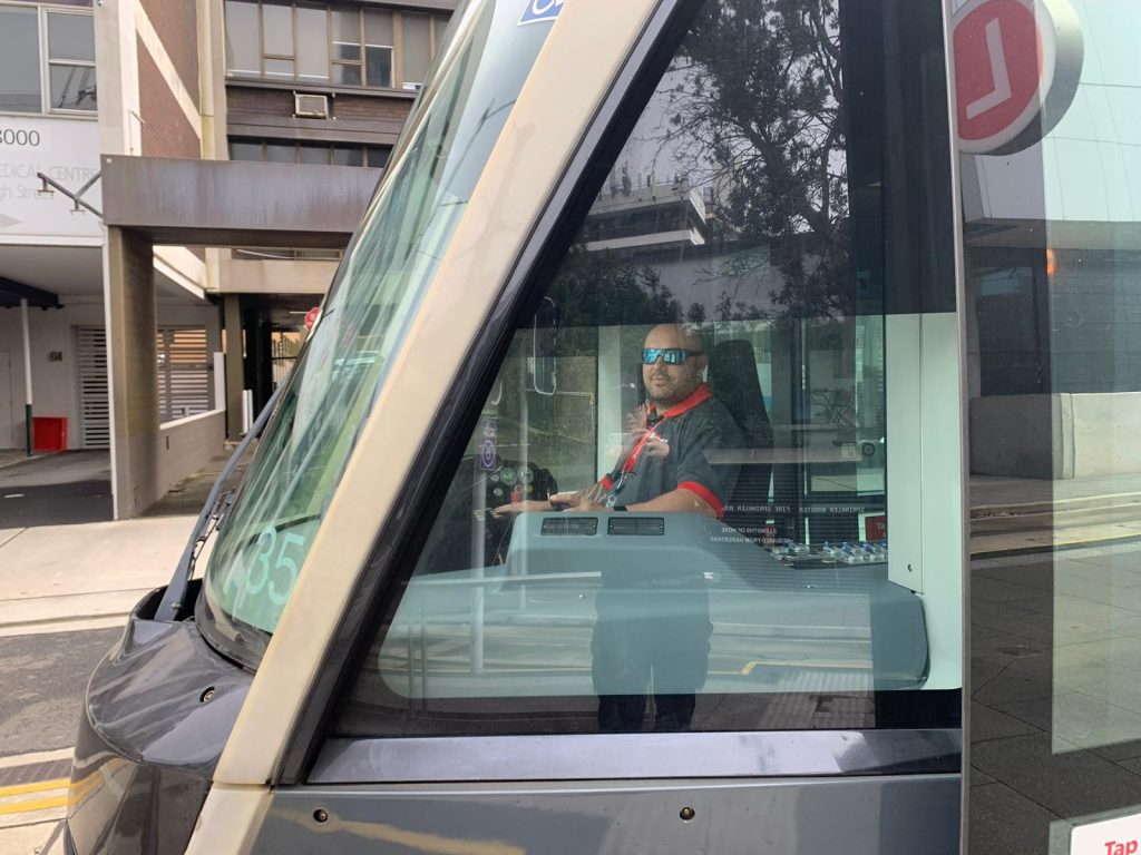 A man in sunglasses looks at the camera from the drivers seat of a light rail vehicle in Sydney's Central Business District.